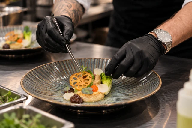 Close-up of gloved hands arranging a dish with tweezers on a ridged plate.